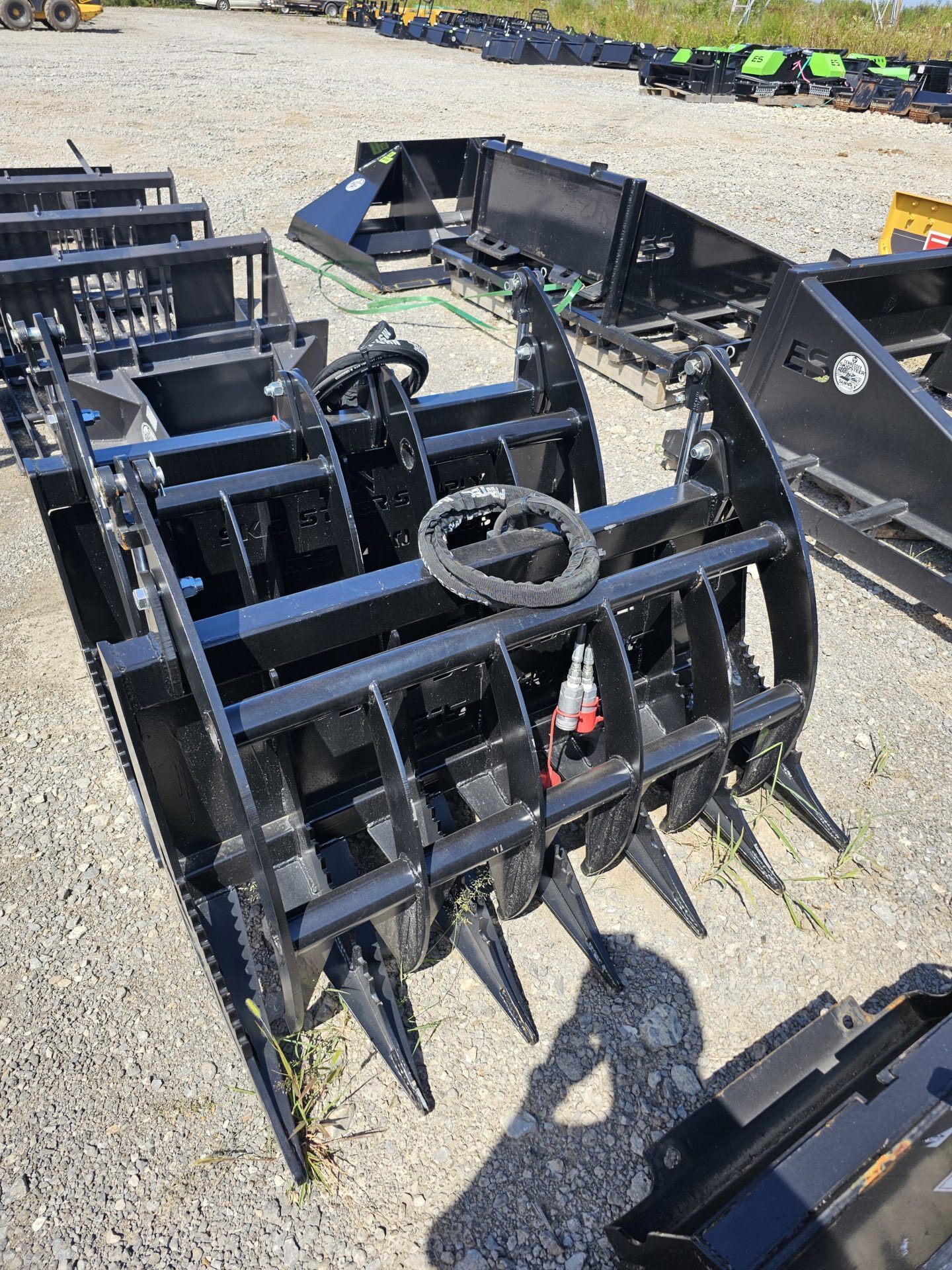 20240821_144722-e1724791807759.jpg The BPT BobcatMT Single/Dual Clam Grapples, a heavy-duty black metal grapple attachment with curved tines and hydraulic components, sits on gravel outdoors amid other equipment for construction or agricultural use.