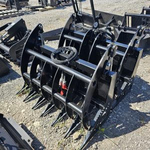 A BPT BobcatMT Single/Dual Clam Grapple, made of black metal, rests on gravel among other equipment, featuring curved tines, hydraulic cylinders, and sharp prongs for heavy machinery.