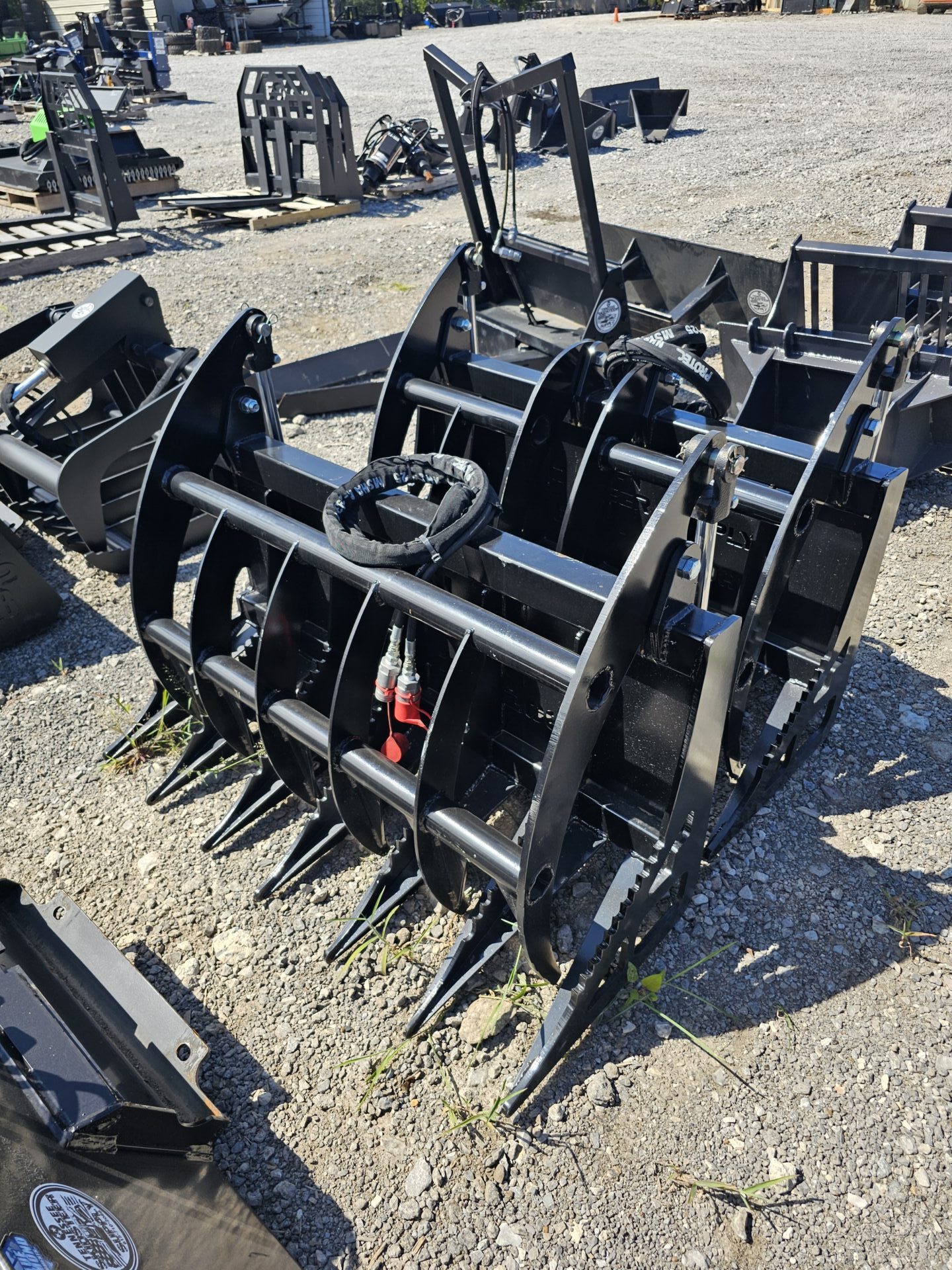 20240821_144727-e1724791794330.jpg A BPT BobcatMT Single/Dual Clam Grapple, made of black metal, rests on gravel among other equipment, featuring curved tines, hydraulic cylinders, and sharp prongs for heavy machinery.