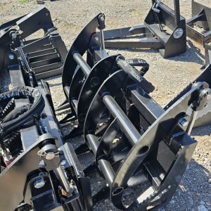 A close-up of BPT BobcatMT Single/Dual Clam Grapples for skid steers, displayed on outdoor gravel among construction equipment. Sunlight emphasizes their heavy-duty black metal build and robust industrial design.