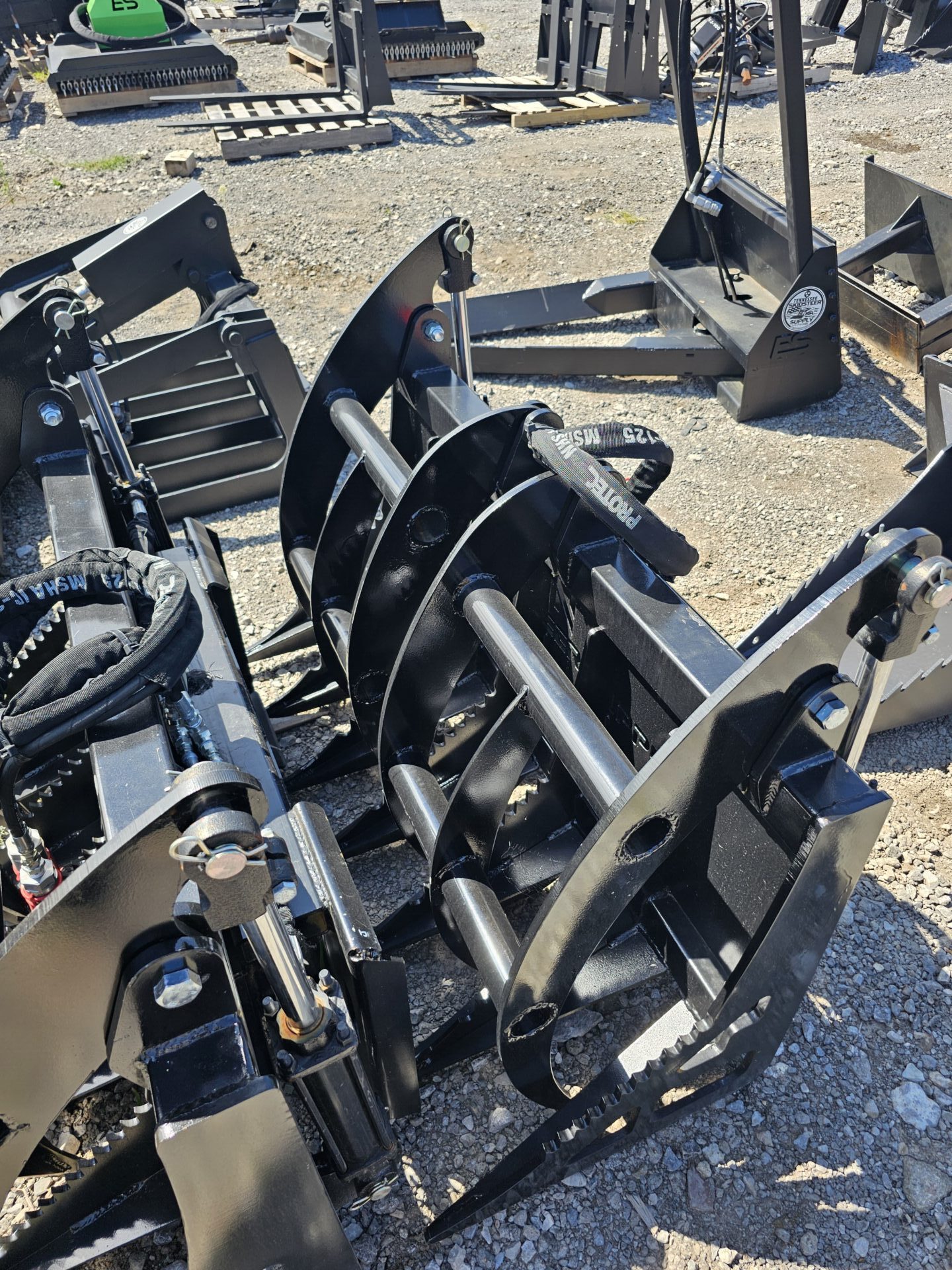 20240821_144731-e1724791770121.jpg A close-up of BPT BobcatMT Single/Dual Clam Grapples for skid steers, displayed on outdoor gravel among construction equipment. Sunlight emphasizes their heavy-duty black metal build and robust industrial design.