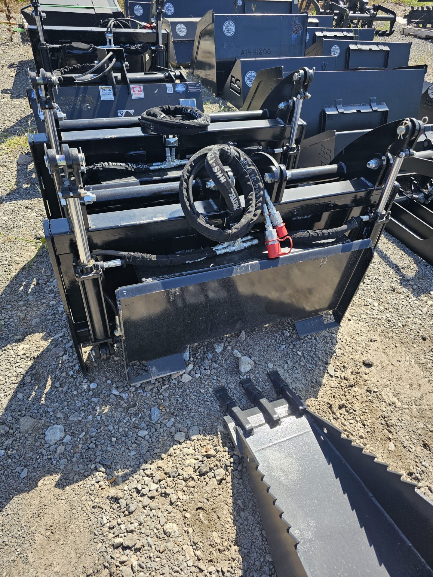 20240821_144735-e1724791733125.jpg A close-up view of several BPT BobcatMT Single/Dual Clam Grapples—heavy-duty black metal attachments for construction equipment—arranged outdoors on gravel, with hydraulic hoses visible on top.