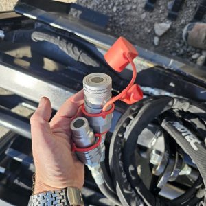 A hand holds two hydraulic quick-connect couplers with red caps beside black hoses and machinery parts outdoors, near a BPT BobcatMT Single/Dual Clam Grapples. Part of a person's boot is visible in the background.