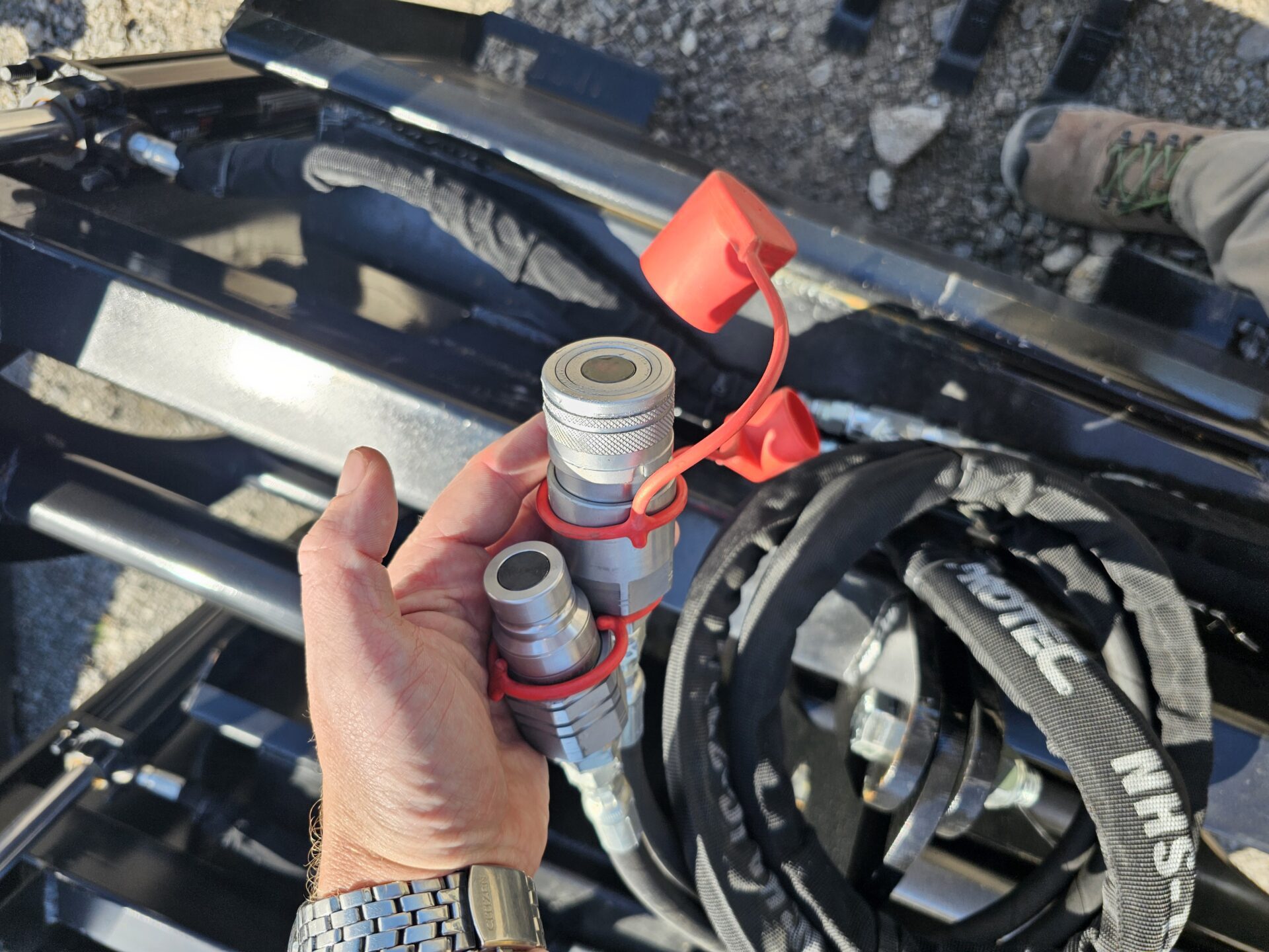 20240821_144802.jpg A hand holds two hydraulic quick-connect couplers with red caps beside black hoses and machinery parts outdoors, near a BPT BobcatMT Single/Dual Clam Grapples. Part of a person's boot is visible in the background.