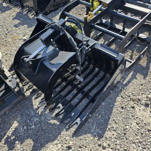 A Prime Cyclops Grapple, made of black metal with curved tines and hydraulic parts, sits on gravel among similar attachments, with a few small green plants growing nearby.