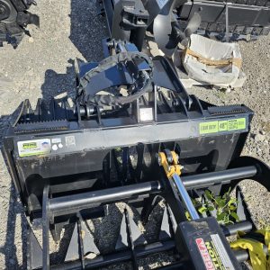A Prime Cyclops Grapple attachment for heavy machinery rests on gravel among other equipment, with its label visible. Some green plants are growing nearby.