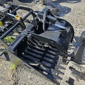 The Prime Cyclops Grapple, a heavy-duty black grapple bucket for construction equipment, rests on gravel among other metal machinery parts and scattered plants.