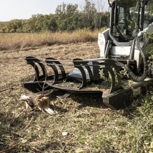 A Prime 80" Direct Drive Brush Cutter is attached to a skid steer loader parked on a field covered in cut branches and debris, with trees and tall grass in the background.