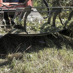 A close-up of the Prime 80" Direct Drive Brush Cutter clearing and shredding brush and vegetation on grassy terrain, with its rotating blades and collected plant debris visible.