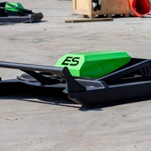 The ES 60" Standard Brush Cutter (up to 60HP), featuring green and black colors with "ES" on top, is positioned on a concrete surface outdoors among various equipment and machinery.