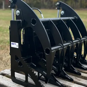 A BPT BobcatMT Single/Dual Clam Grapple in black metal with curved tines sits on a wooden pallet outdoors on grass, trees in the background, and a visible BPT label.