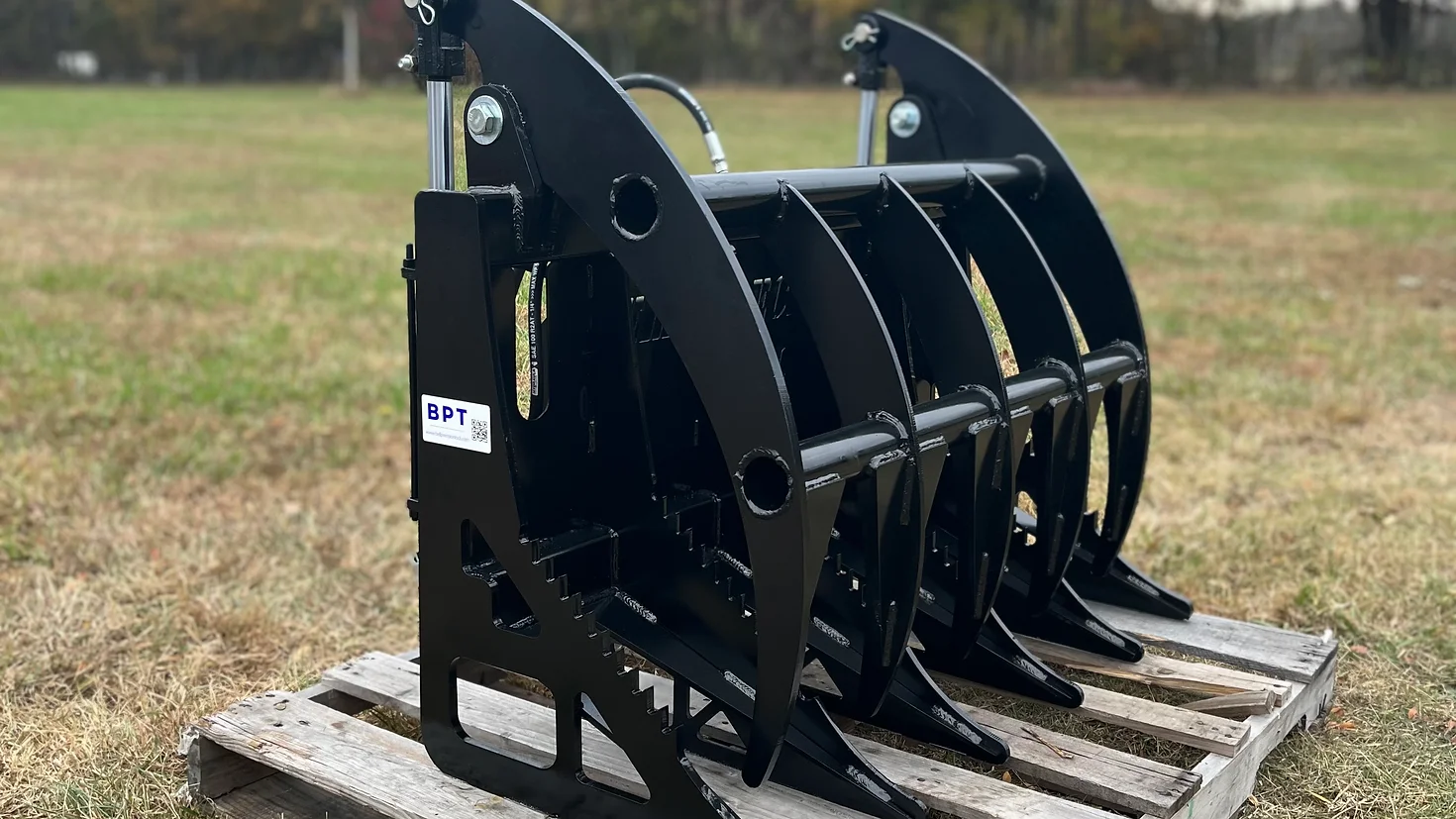 ce79f6_d59a609970be42c680f93c19cfba6187mv2-2.webp A BPT BobcatMT Single/Dual Clam Grapple in black metal with curved tines sits on a wooden pallet outdoors on grass, trees in the background, and a visible BPT label.